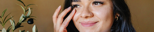 A woman gently applying skincare under her eyes, smiling with a radiant complexion, set against a warm, natural background.