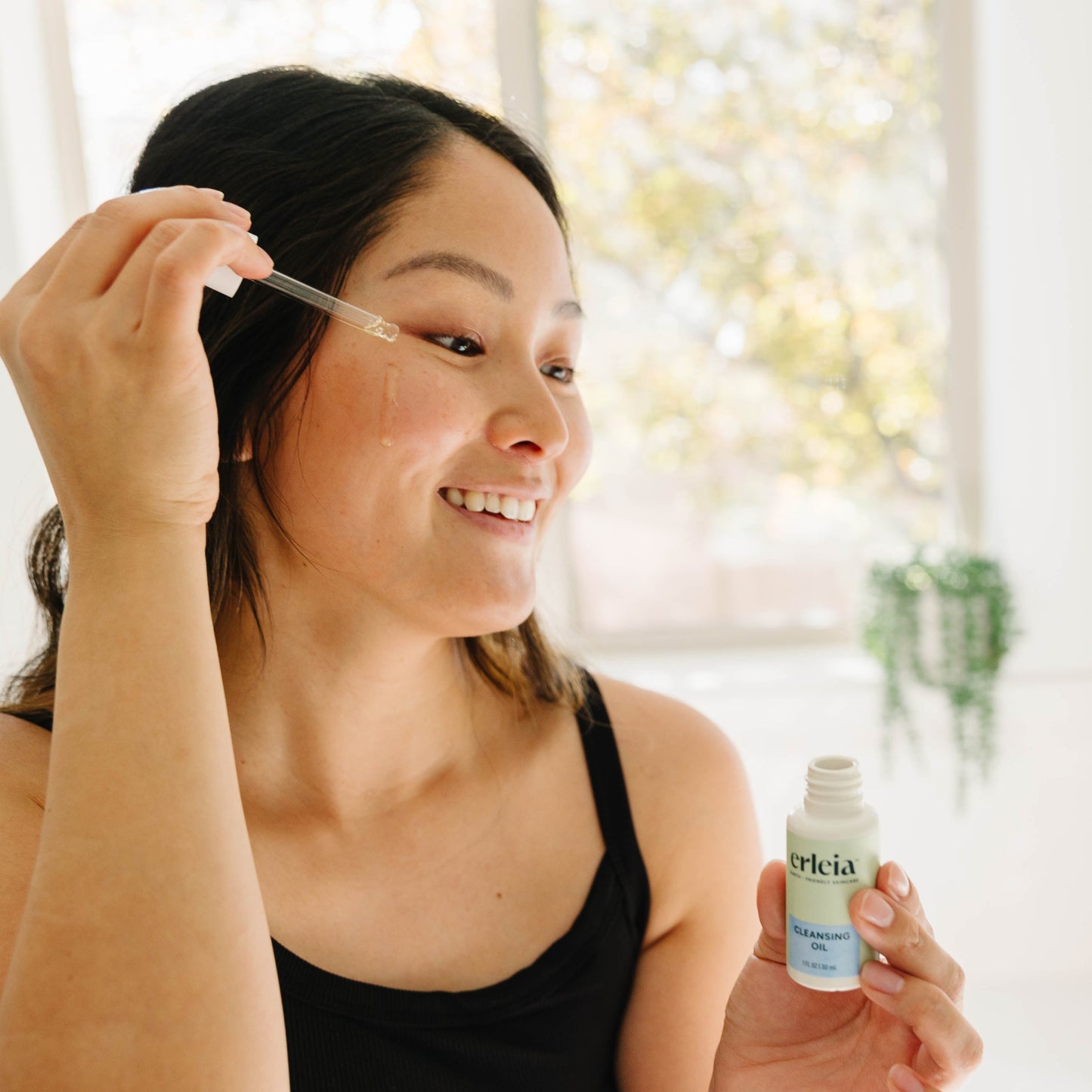 An East Asian woman applying Erleia Cleansing Oil with the dropper to her cheek