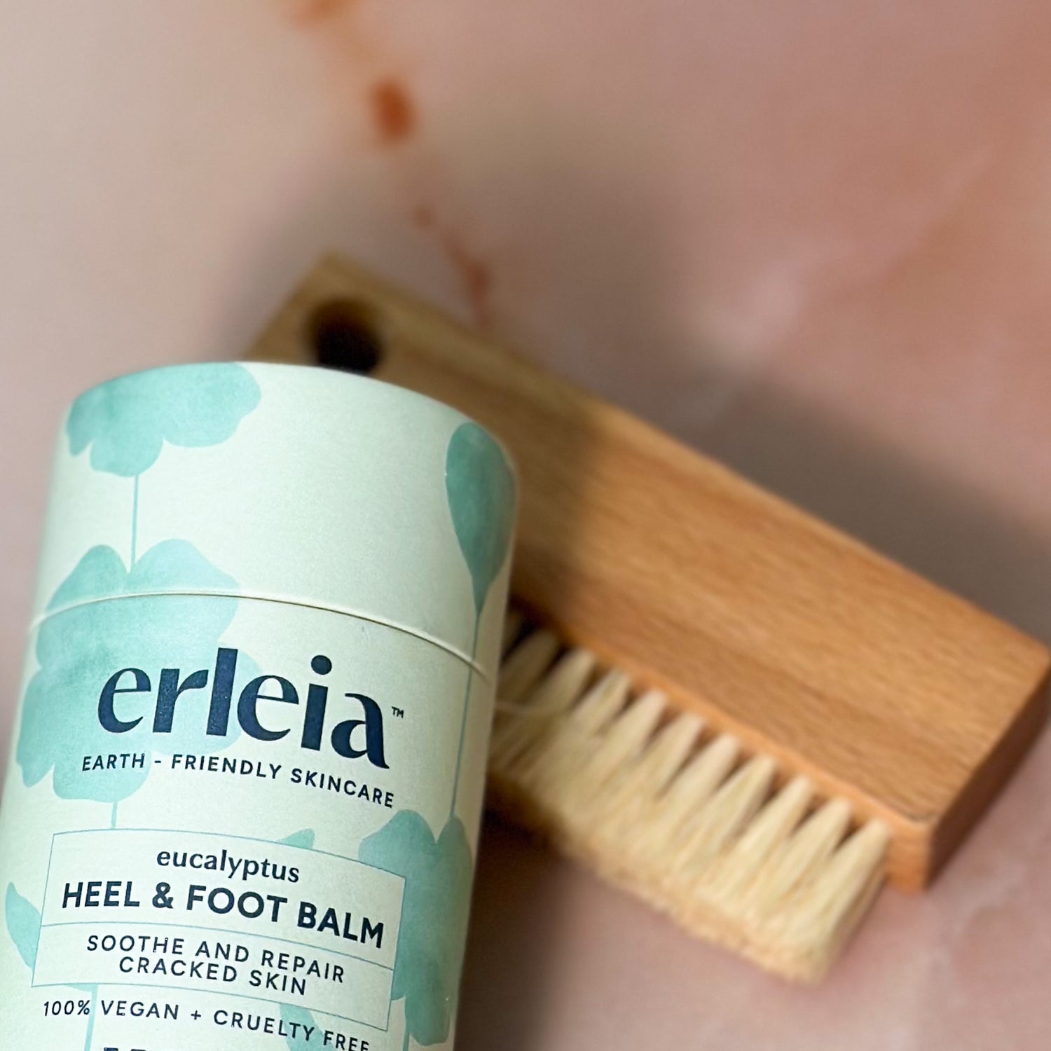 Erleia Eucalyptus Heel & Foot Balm is displayed in front of a wooden foot brush with a soft pink background.