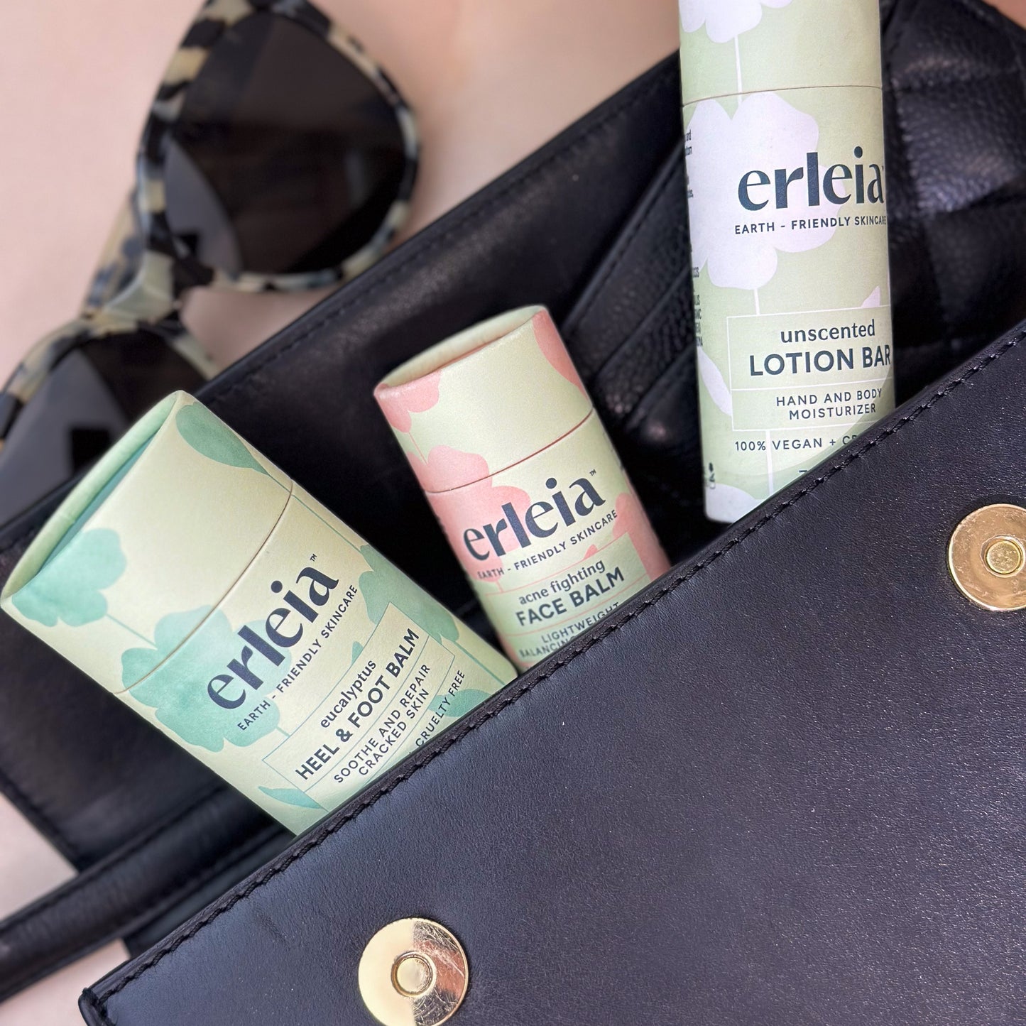 A black bag is open to reveal Erleia skincare products including a heel and foot balm, face balm, and lotion bar, with a pair of sunglasses placed beside the bag.