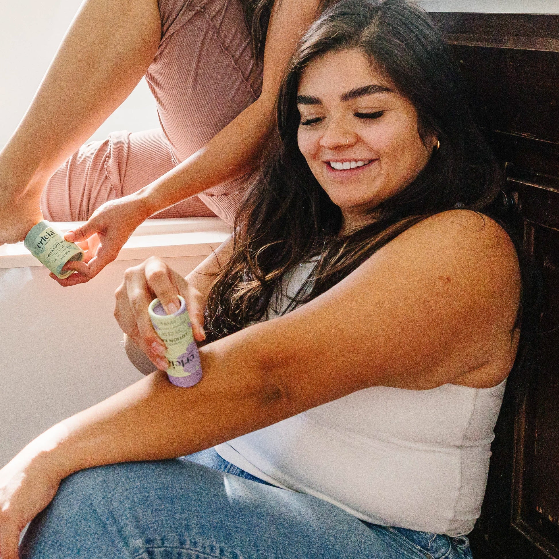 A light skinned Latina woman applying an Erleia Lavender Lotion Bar to her arm while sitting in a bathroom