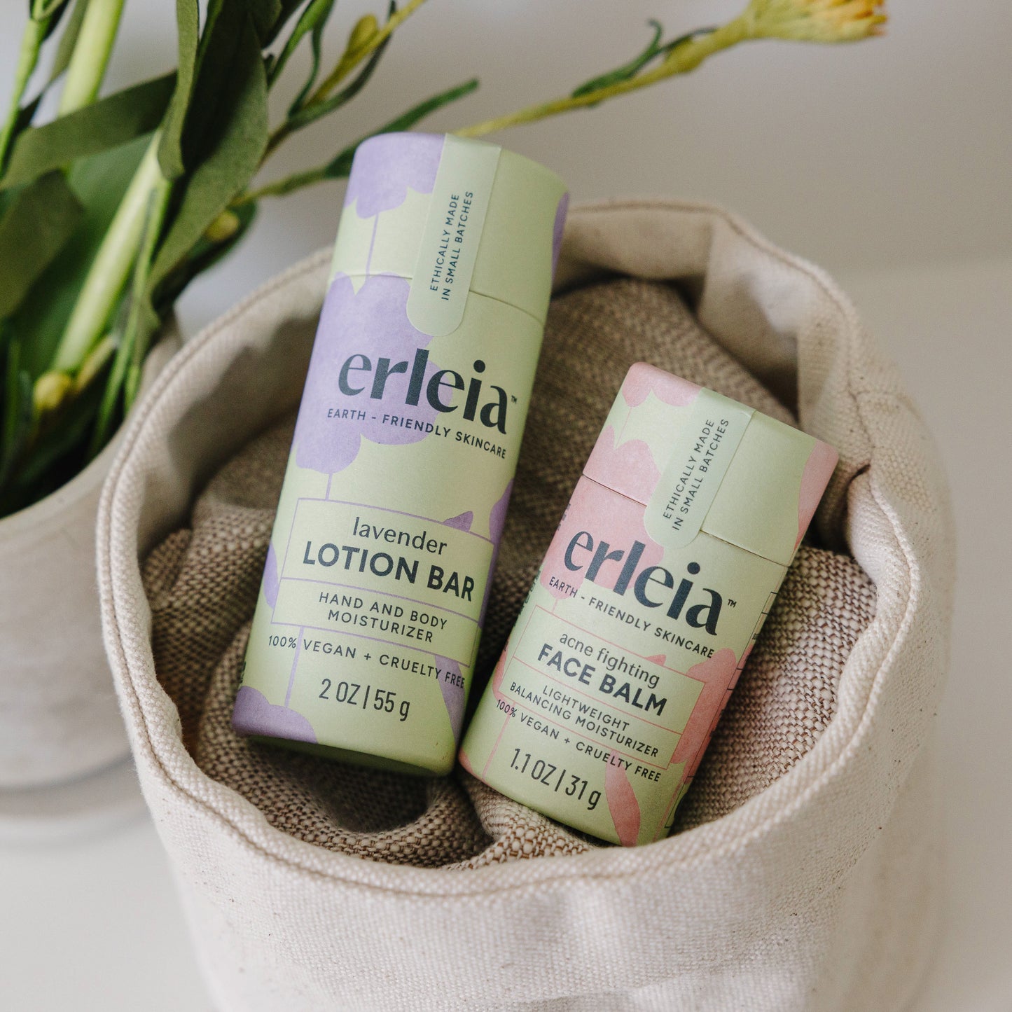 Erleia lavender lotion bar and acne-fighting face balm are displayed in eco-friendly packaging, resting inside a beige fabric basket with a plant in the background.