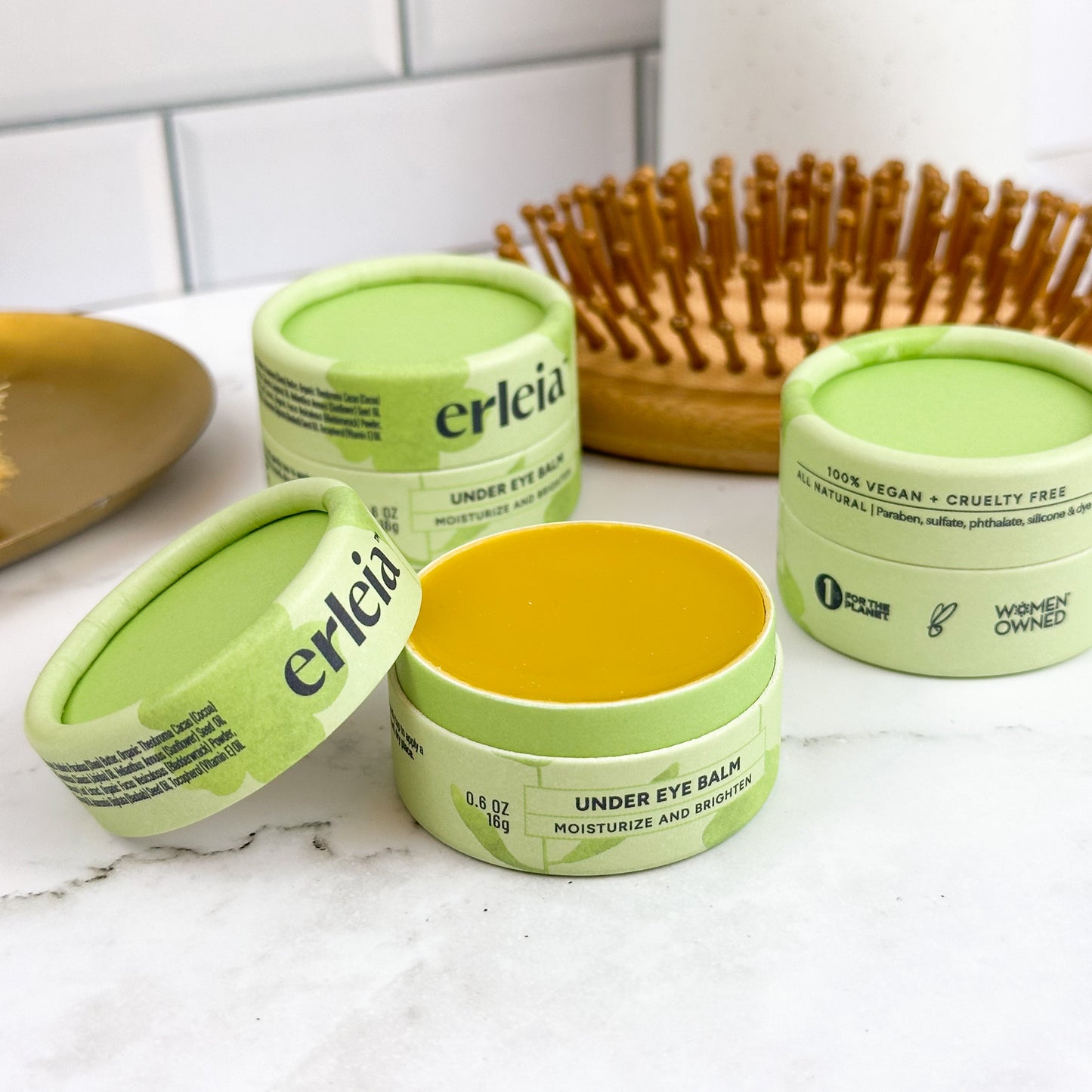 Three open jars of Erleia Under Eye Balm are displayed on a marble surface alongside a wooden hairbrush, with one jar showing the smooth balm inside. The packaging highlights the product as vegan, cruelty-free, and women-owned.