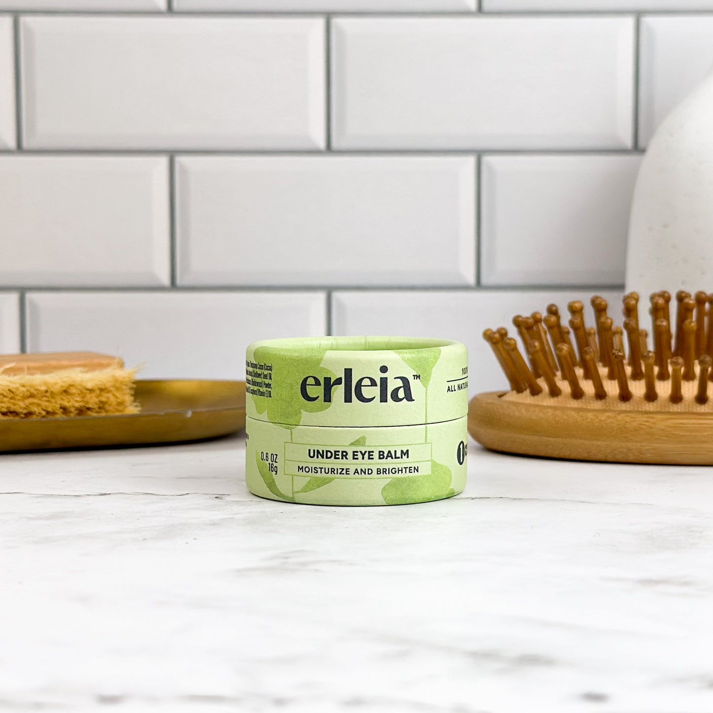 A jar of Erleia Under Eye Balm is displayed on a marble surface, with a wooden hairbrush and skincare accessories in the background. The product packaging highlights its benefits for moisturizing and brightening the under-eye area.
