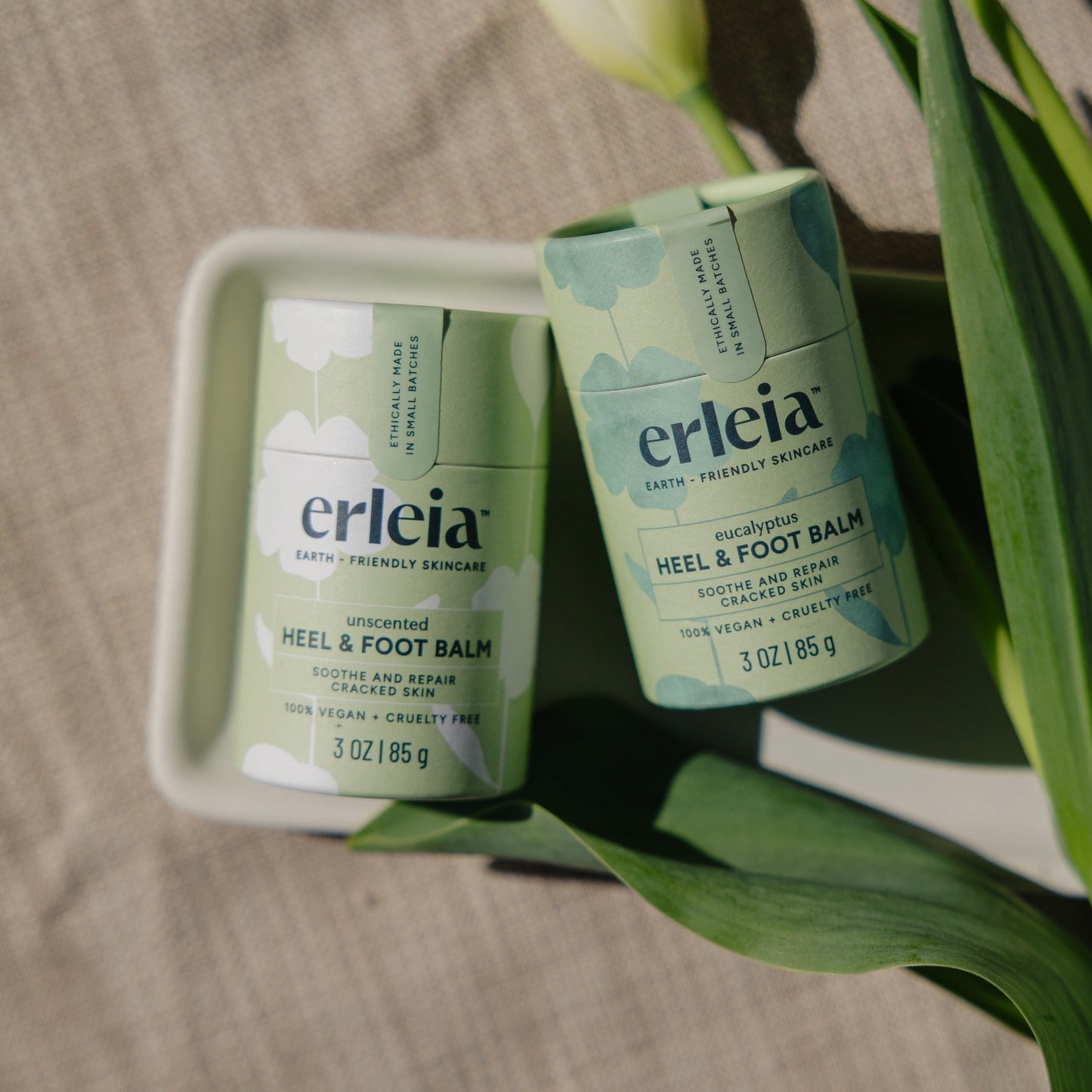 Two Erleia Heel & Foot Balms, one unscented and the other eucalyptus, are displayed in eco-friendly packaging with tulip leaves surrounding them.