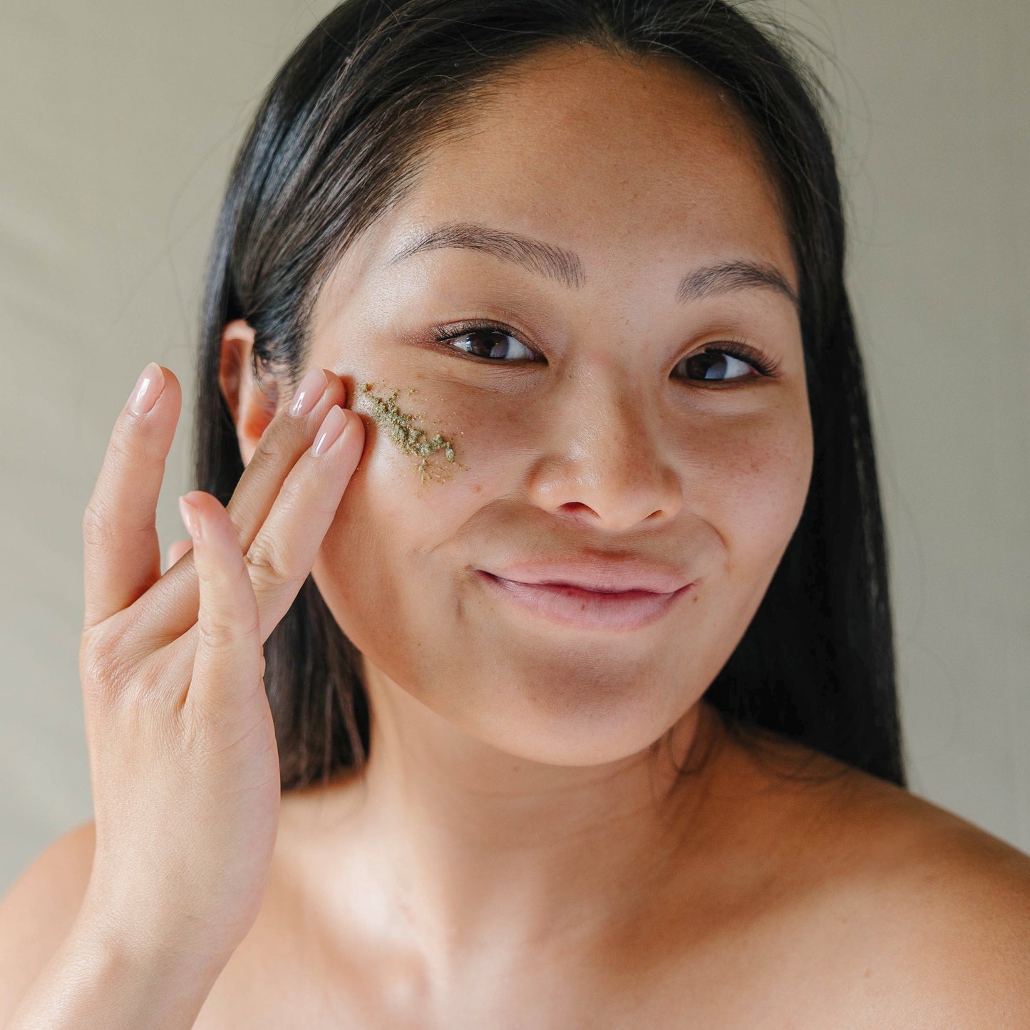 An East Asian woman smiling and applying Erleia Humble Powder Cleanser, a green powder, to her cheek