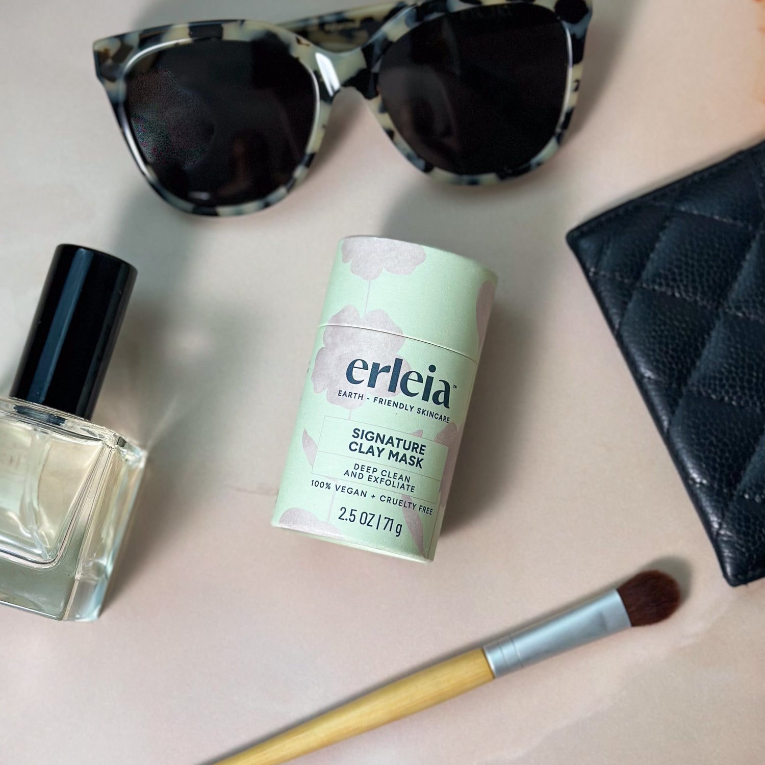 A flat lay of beauty essentials, featuring Erleia's Signature Clay Mask, sunglasses, a makeup brush, a perfume bottle, and a black quilted pouch.