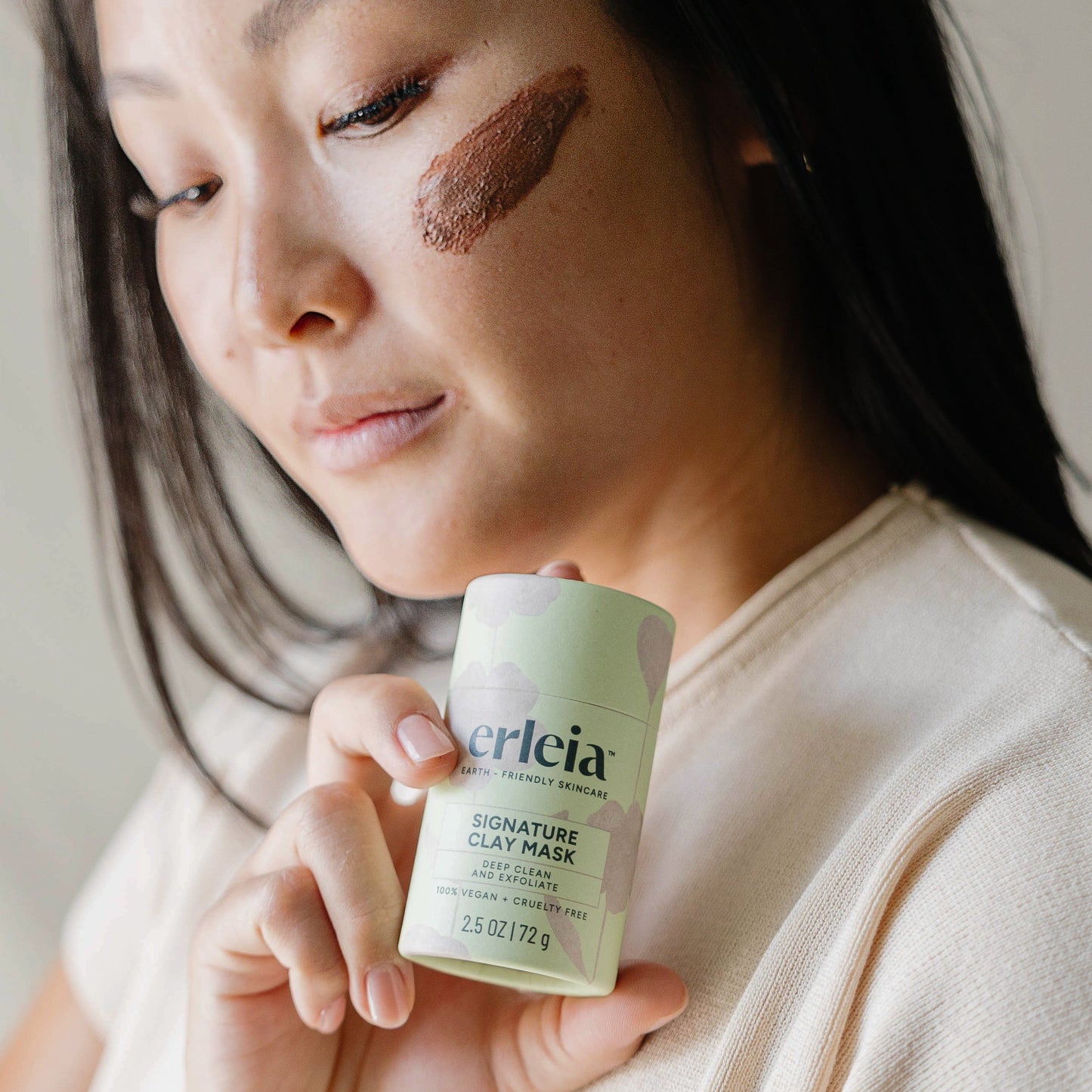 An East Asian woman holding an Erleia Signature Clay Mask, she has some of the mask applied to her cheek