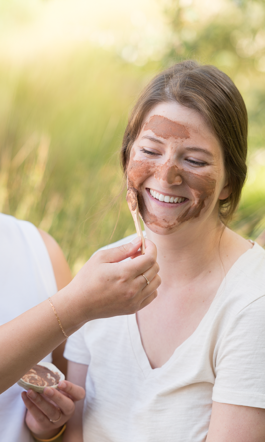 An East Asian hand applying a clay mask with a spoon to a white woman