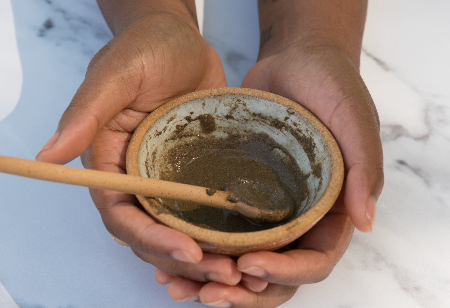 Hands are holding a small ceramic bowl containing the Erleia Humble Powder Cleanser, with a wooden mixing stick resting inside the bowl.