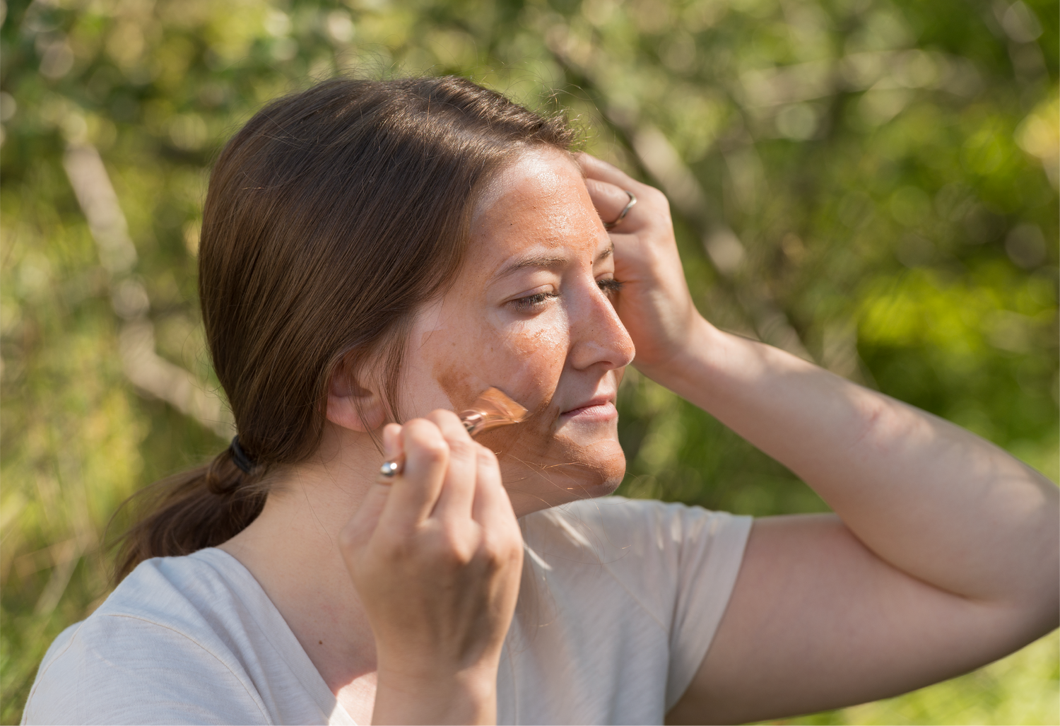 A woman is applying the Erleia Signature Clay Mask to her face with a brush in an outdoor setting, highlighting a natural and serene moment.