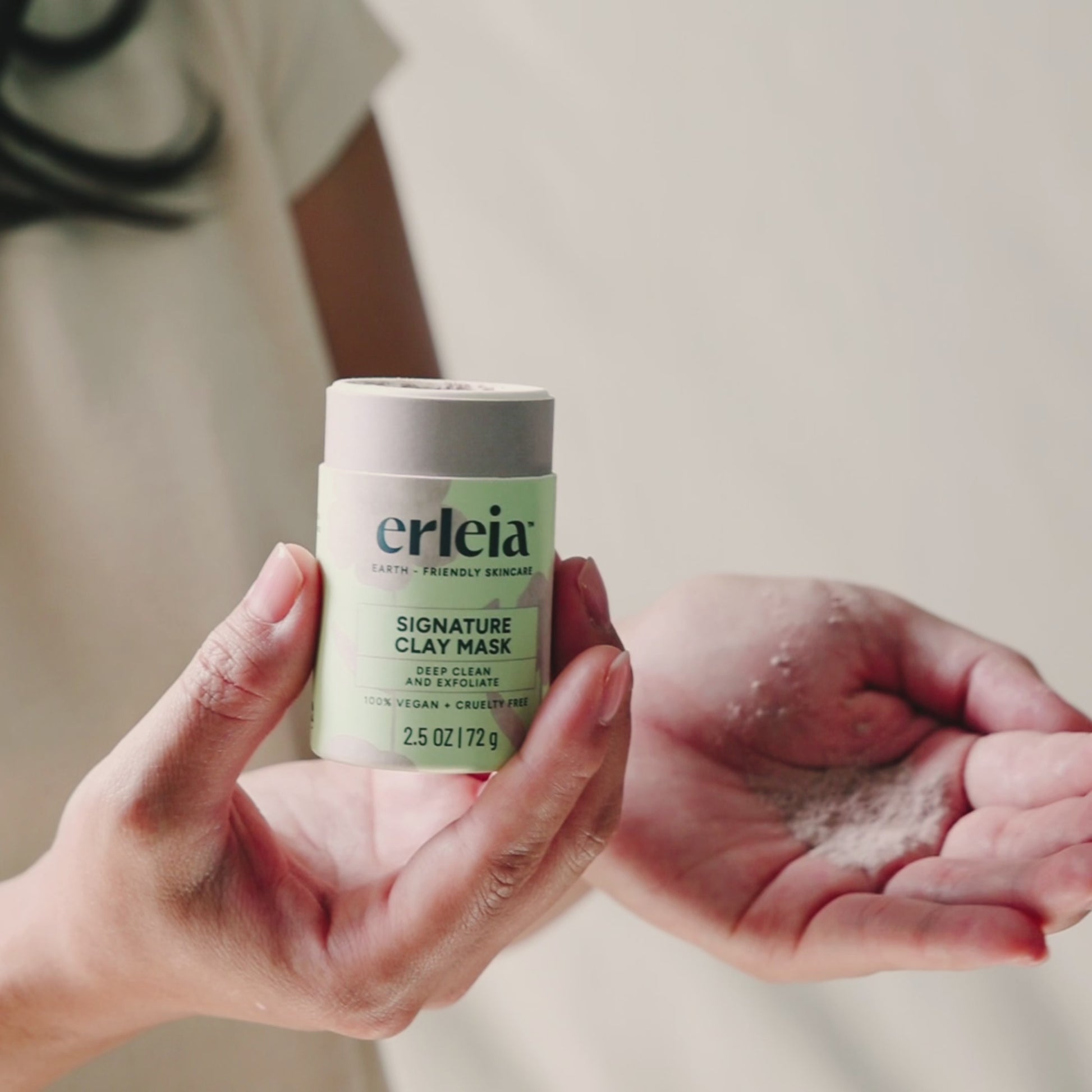 An East Asian woman pours Erleia Signature Clay Mask from the packaging into her hand and then applies it to a Black Woman's face, they both have soft smiles.