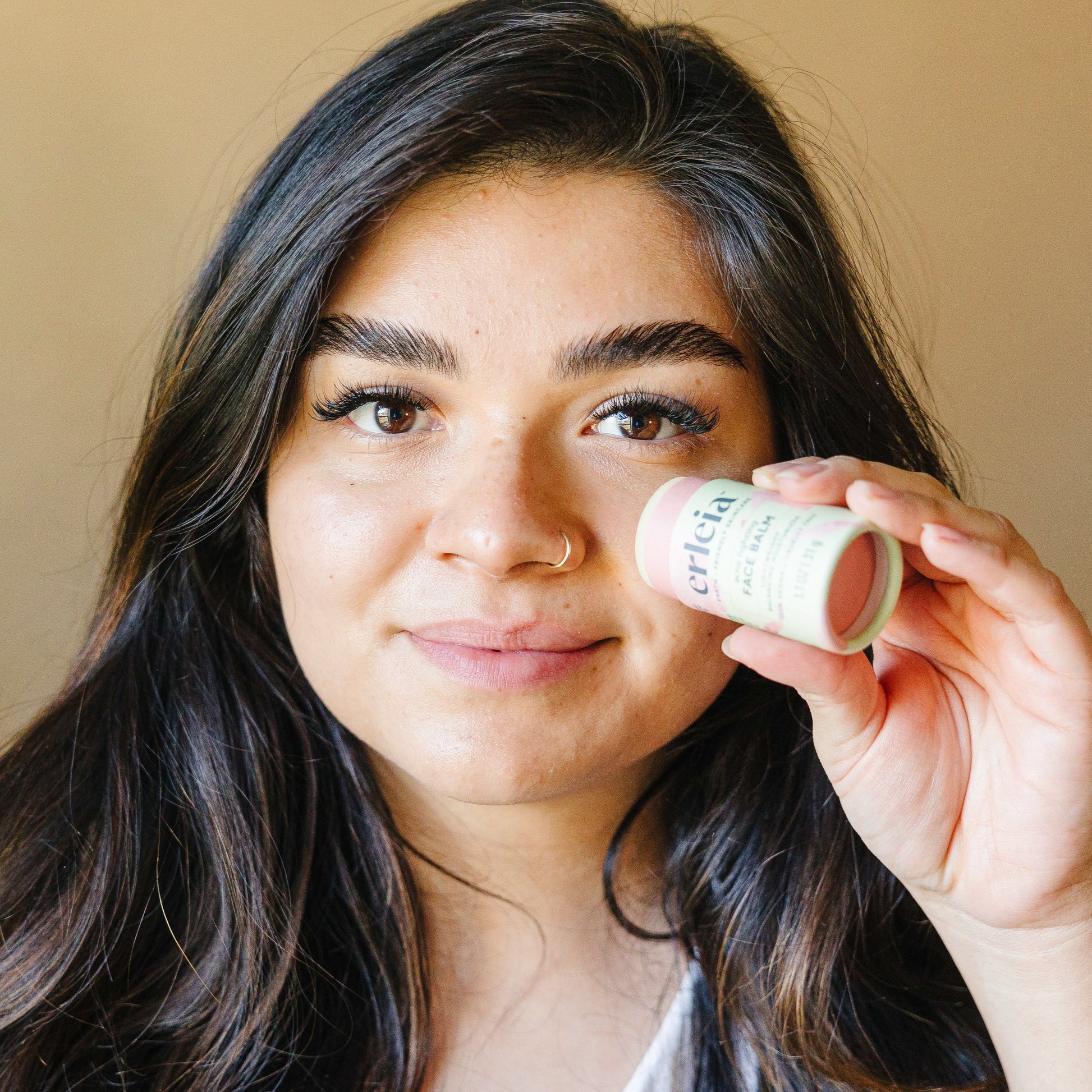 An light skinned Latina woman holding an Erleia Acne Fighting Face Balm and applying it to her cheek while looking at the camera