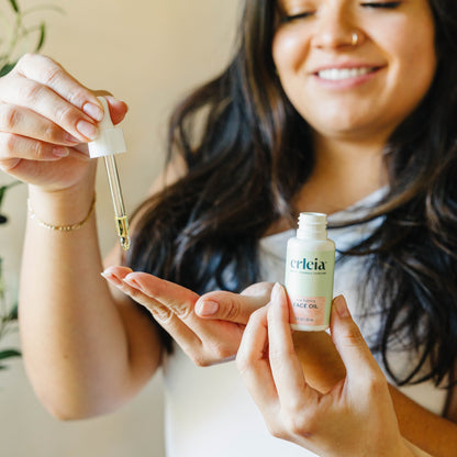 A light skinned Latina woman using a dropper of Erleia Acne Fighting Face Oil and dropping product on her fingers while another hand holds the bottle