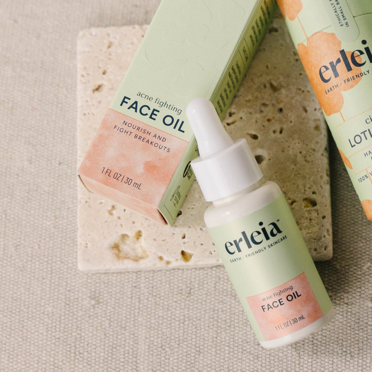 Erleia Acne Fighting Face Oil bottle and box next to an Erleia Citrus Lotion Bar on a neutral textured background