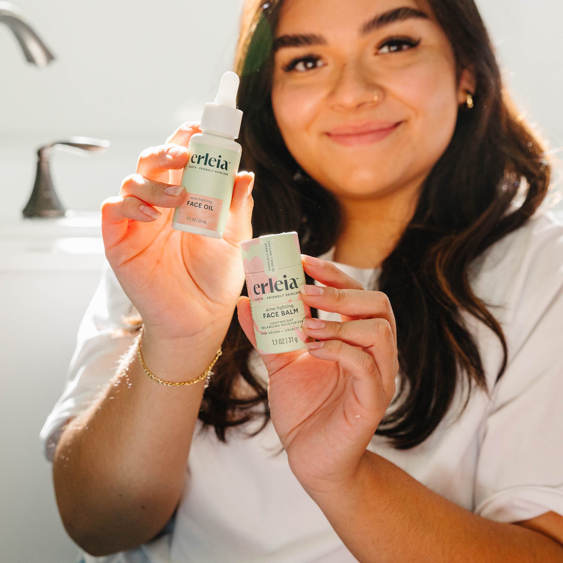 A light skinned Latina woman holding an Erleia Acne Fighting Face Oil and Acne Fighting Face Balm, as she sits in a bathroom