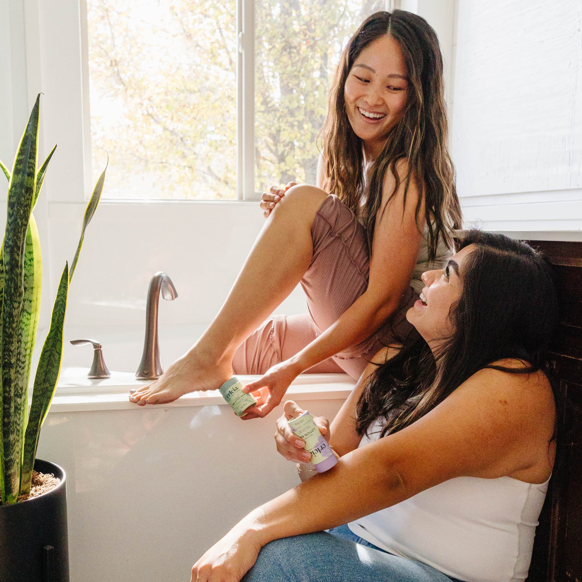 An East Asian woman applies Erleia Heel and Foot Balm to her heel while sitting on a bathtub ledge and smiling at a Latina woman sitting on the ground and applying an Erleia lotion bar to her arm 