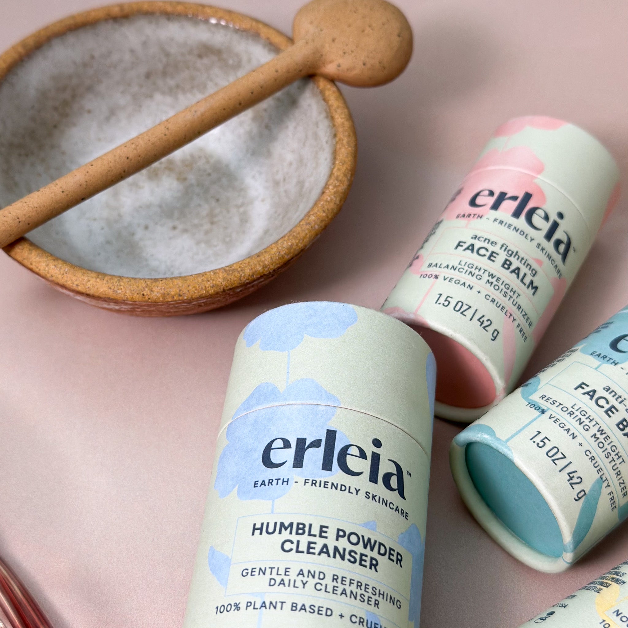 Erleia Humble Powder Cleanser and Face Balm products are displayed next to a ceramic bowl and wooden spoon, creating a soothing skincare setting.