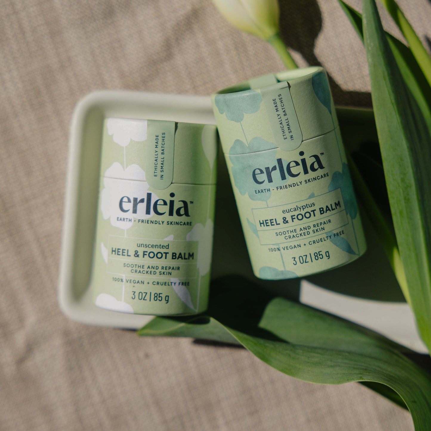 Two Erleia Heel & Foot Balms, one unscented and the other eucalyptus, are displayed in eco-friendly packaging with tulip leaves surrounding them. 