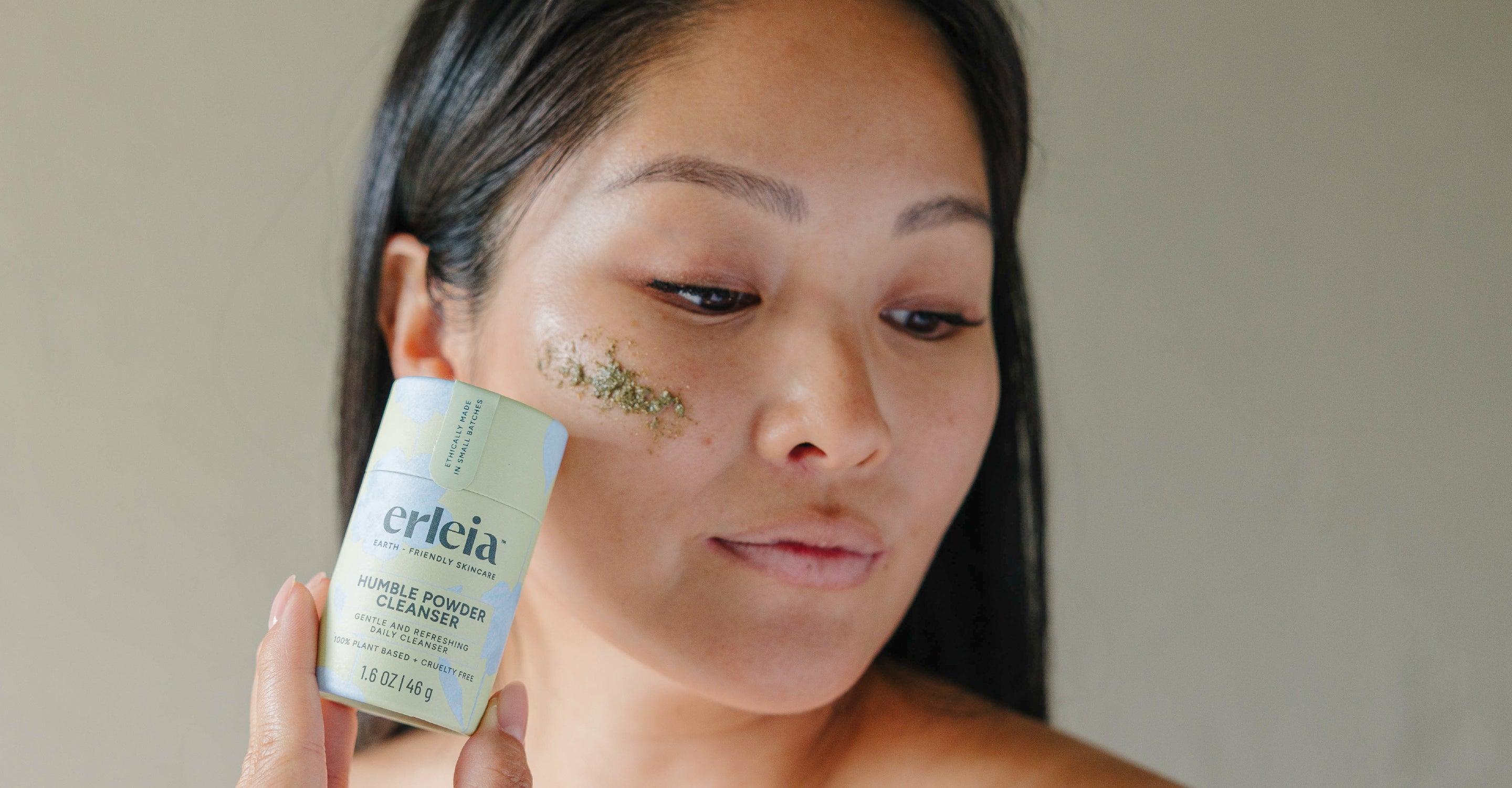 An Asian woman holding an Erleia Humble Powder Cleanser, she has some green cleanser on her cheekbone