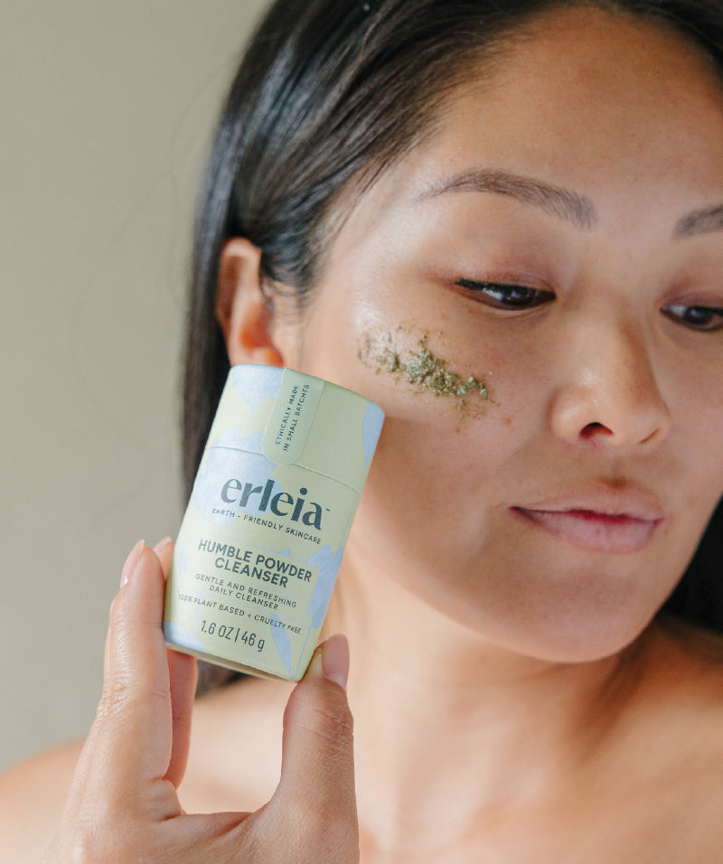 An Asian woman holding an Erleia Humble Powder Cleanser, she has some green cleanser on her cheekbone
