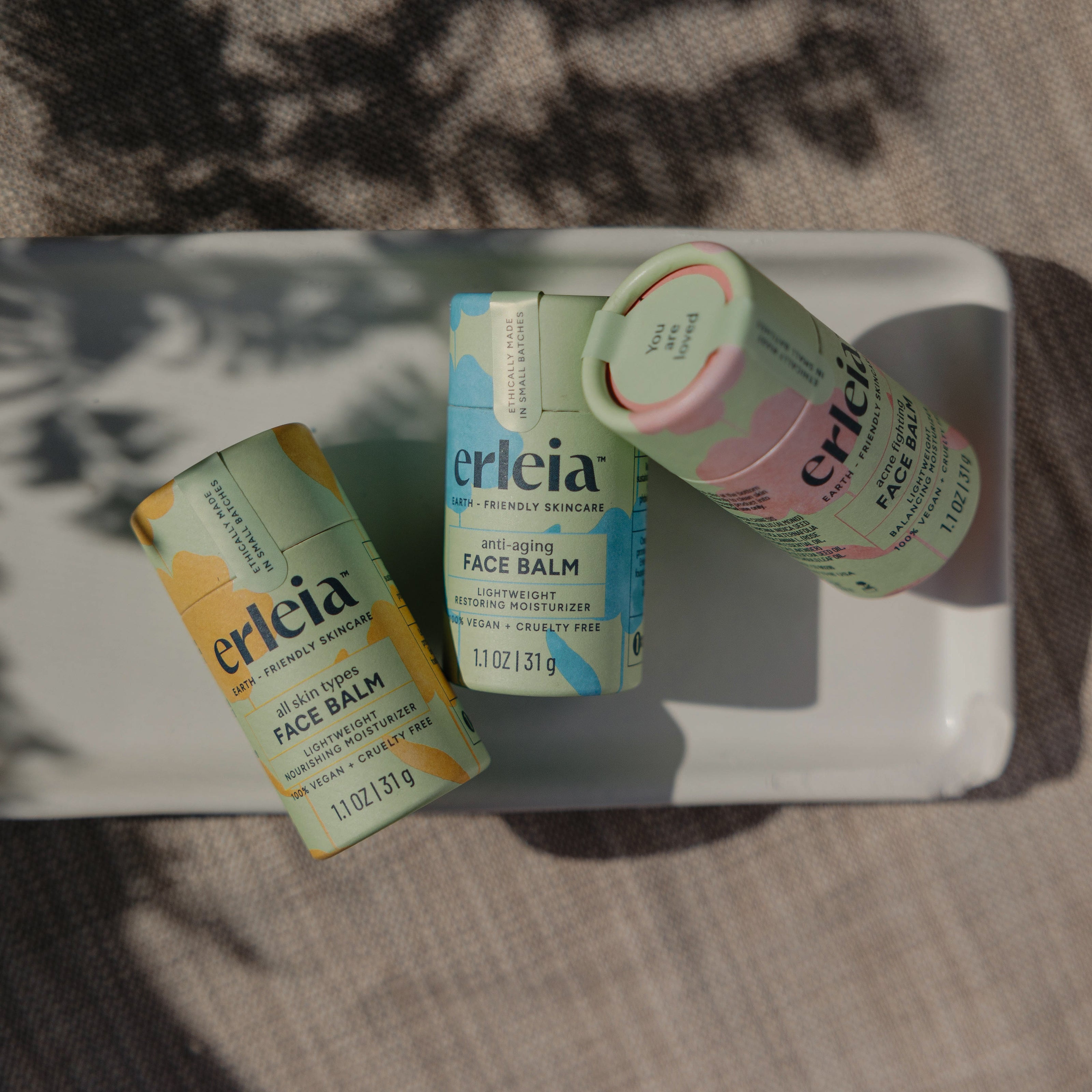 Three Erleia face balms, including anti-aging and all-skin types, are arranged on a white dish, showcasing their earth-friendly packaging with shadows cast by nearby foliage.