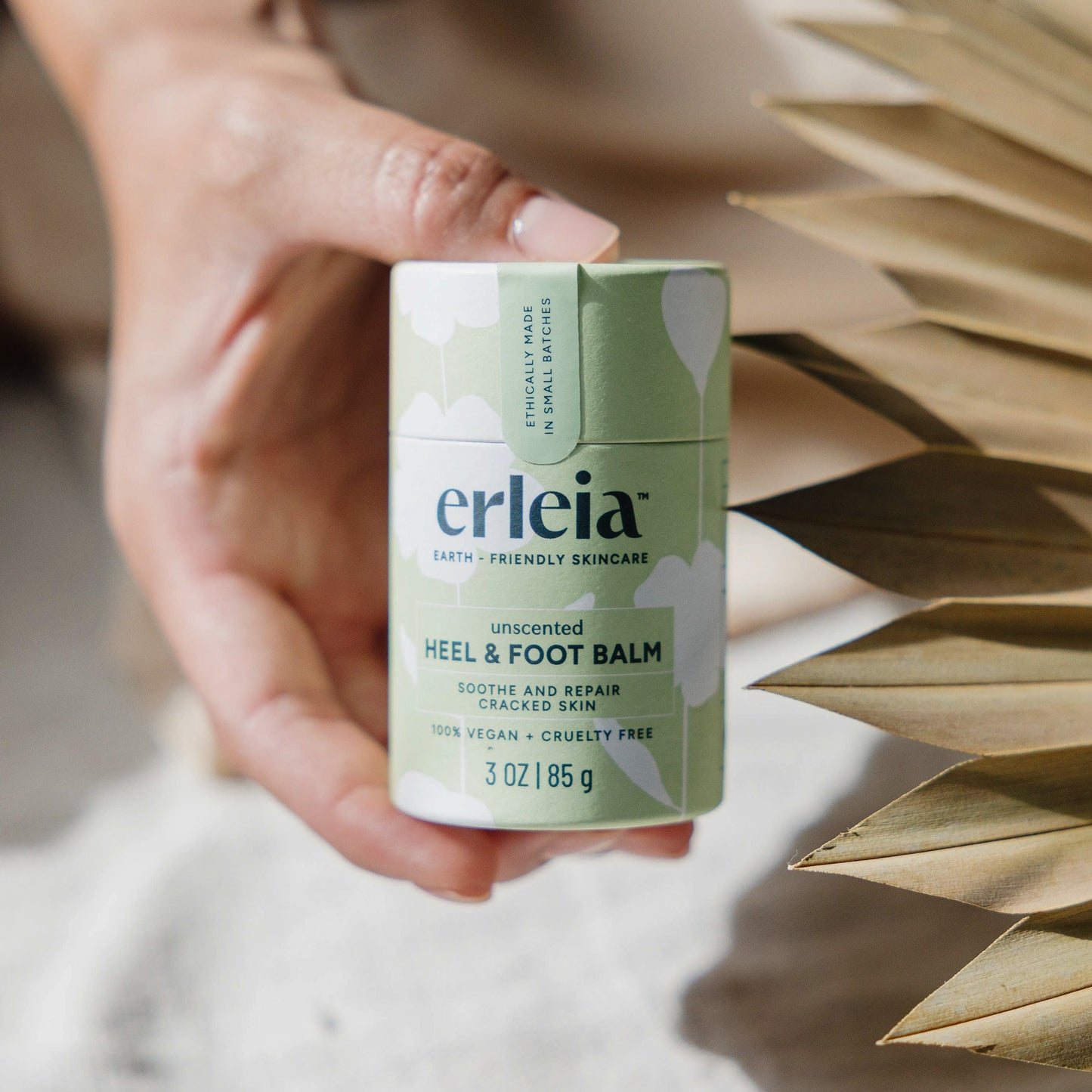 An Erleia unscented Heel & Foot Balm held by an East Asian hand by some dried florals