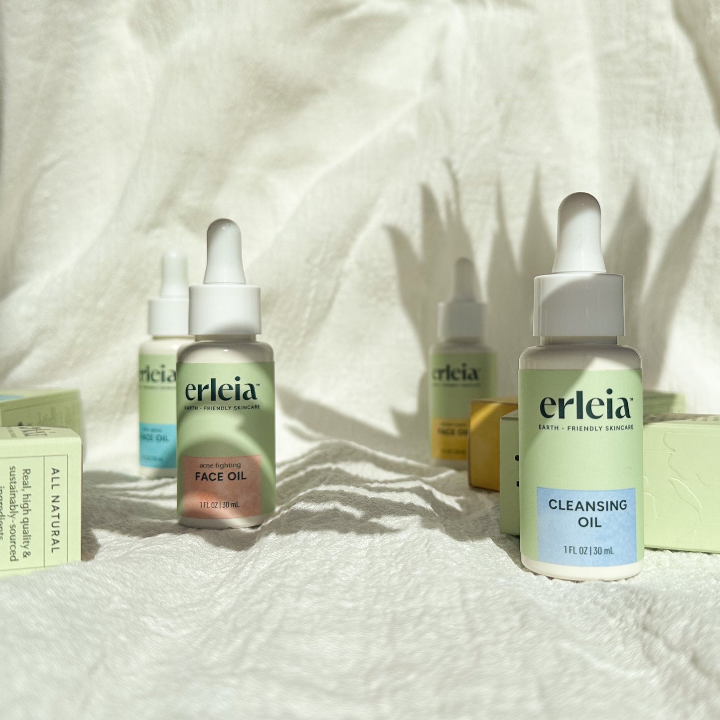 Erleia Oils on a White Fabric Textured Background - there are 3 face oils in different formulas, with bottles and boxes visible, and a Cleansing Oil at front of the photo