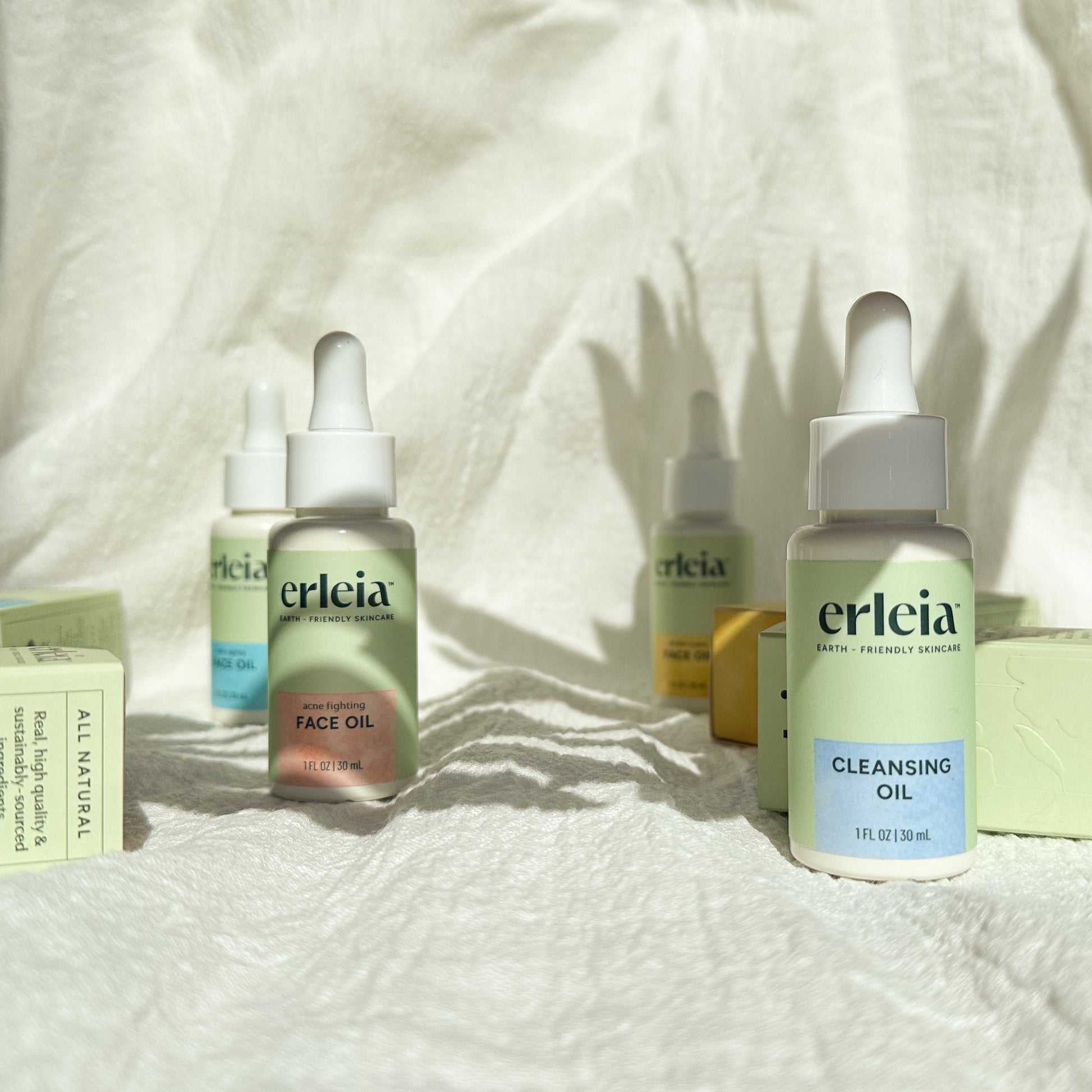 Erleia Oils on a White Fabric Textured Background - there are 3 face oils in different formulas, with bottles and boxes visible, and a Cleansing Oil at front of the photo