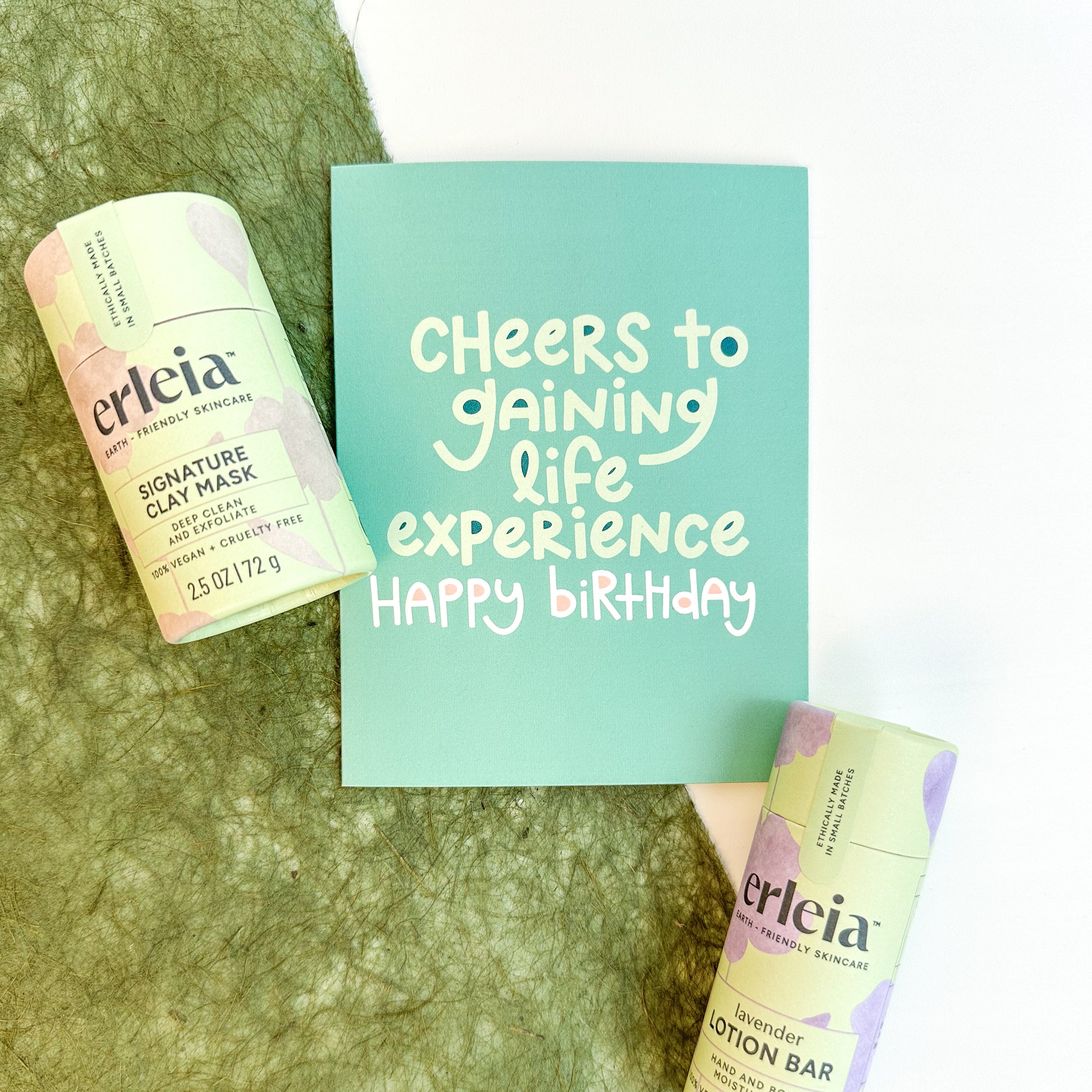 Erleia’s signature clay mask and lavender lotion bar are displayed alongside a birthday card that reads "Cheers to gaining life experience, happy birthday," emphasizing celebration and self-care.