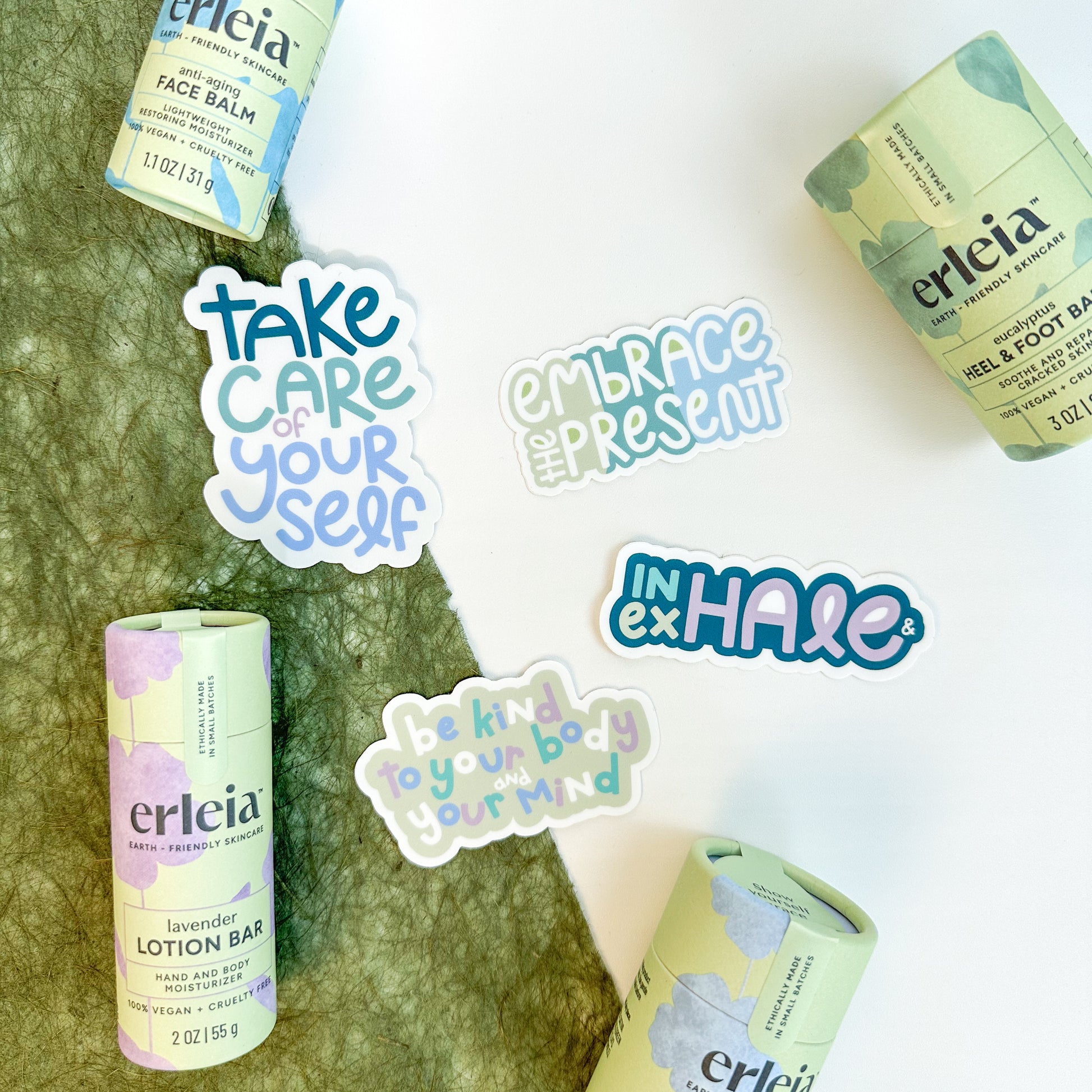 Erleia skincare products, including face balm, heel & foot balm, and lavender lotion bar, are arranged around stickers with messages like "Take care of yourself," "Embrace the present," and "Be kind to your body and your mind," promoting self-care and mindfulness.