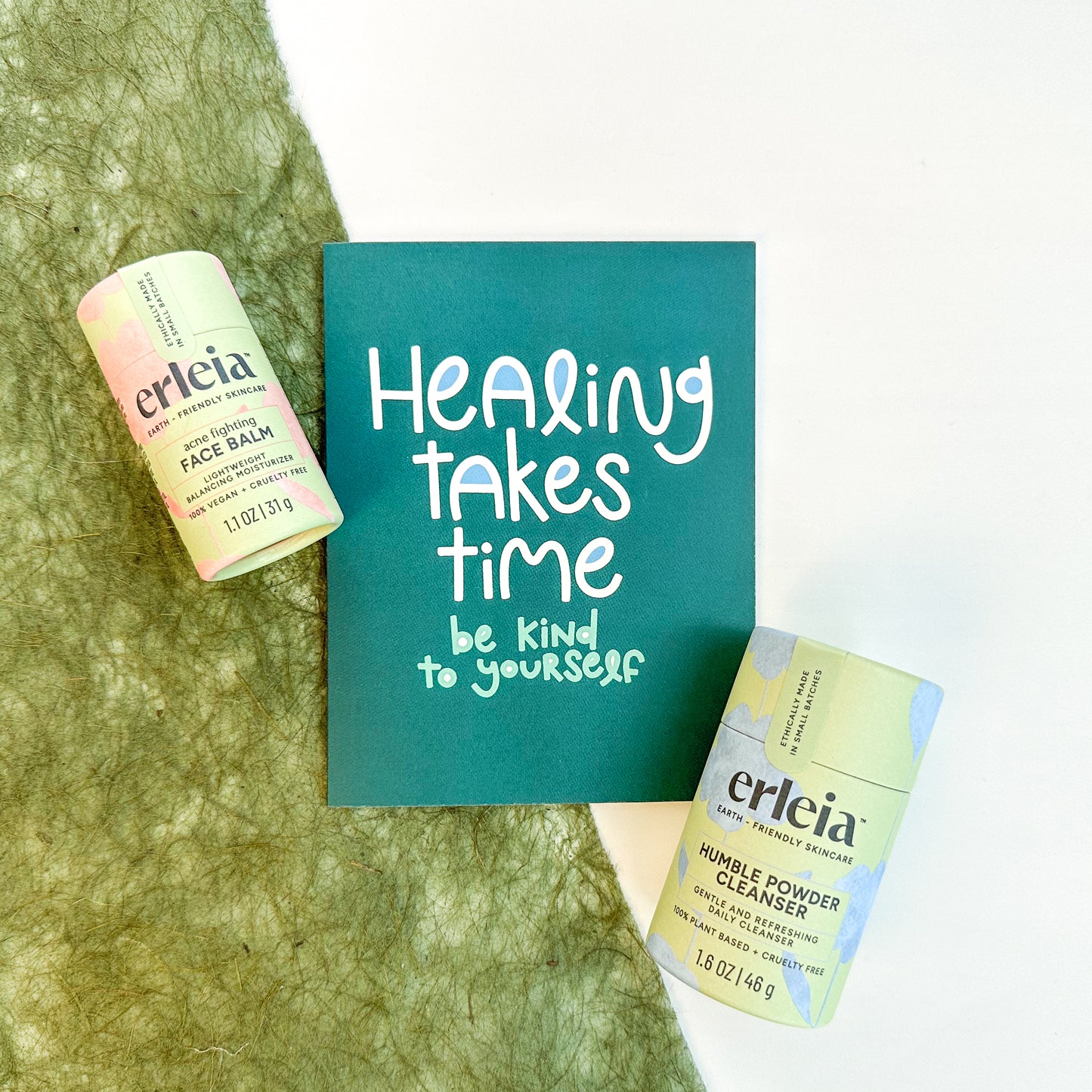 Erleia acne-fighting face balm and humble powder cleanser are displayed alongside a card that reads "Healing takes time, be kind to yourself," promoting self-care and patience in the healing process.