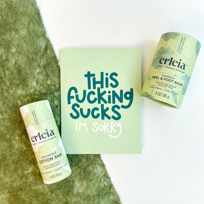 Erleia unscented lotion bar and eucalyptus heel & foot balm are displayed alongside a card that reads "This fucking sucks. I'm sorry," emphasizing empathy and care.