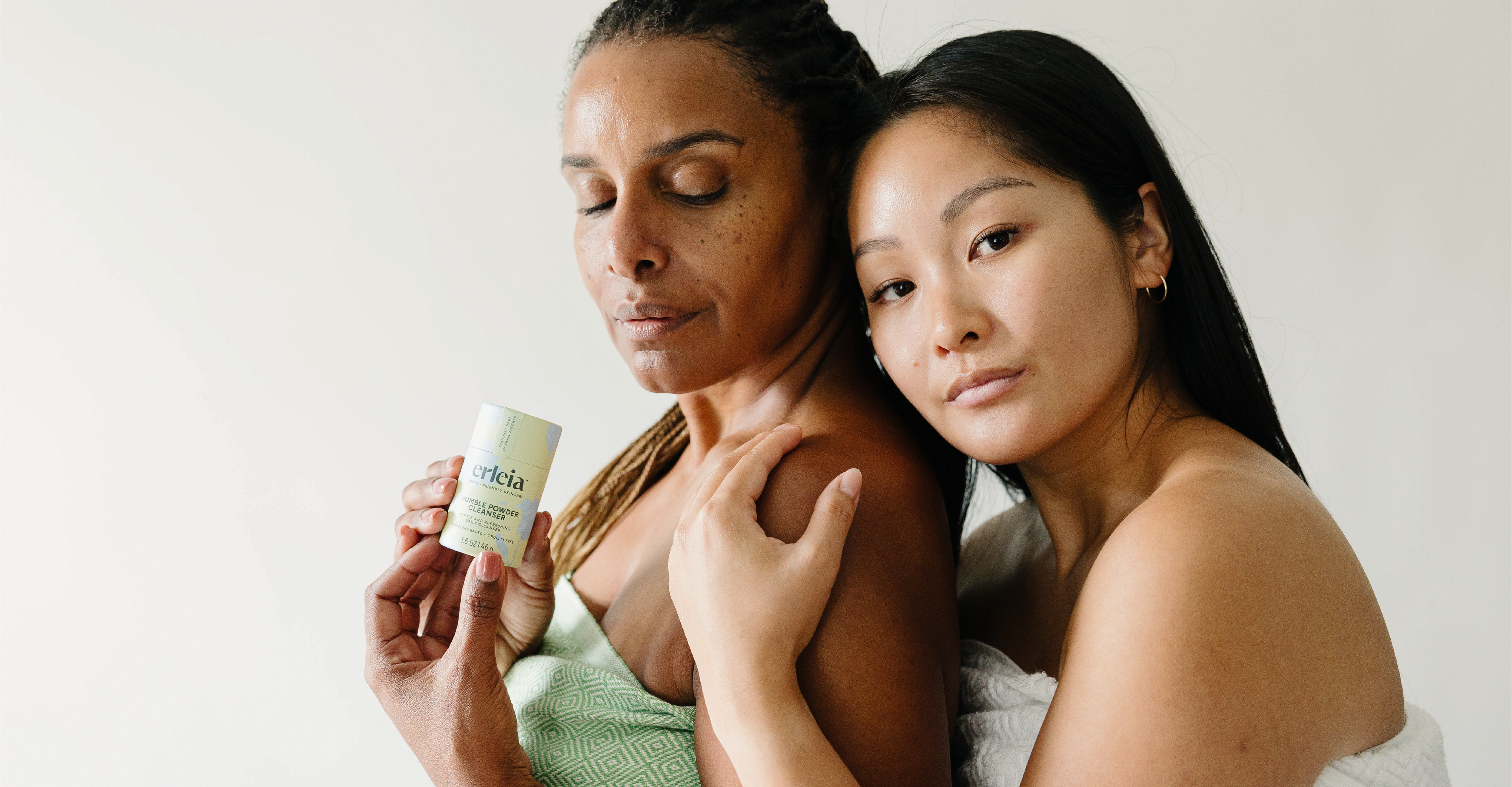 A Black woman and East Asian woman wrapped in towels stand close together, one holding Erleia’s Humble Powder Cleanser, both with calm expressions and glowing skin, emphasizing natural beauty and self-care.