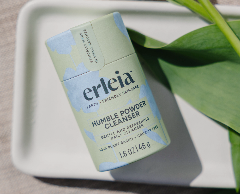 Erleia’s Humble Powder Cleanser is displayed on a white tray in green and blue packaging, emphasizing its gentle, refreshing daily cleansing benefits and 100% plant-based, cruelty-free formula.