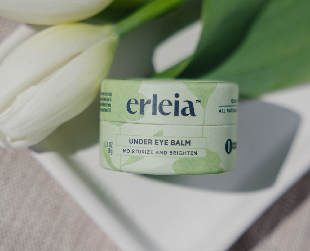 Erleia’s under eye balm is displayed in eco-friendly packaging alongside white tulips, highlighting its moisturizing and brightening benefits with an all-natural formula.