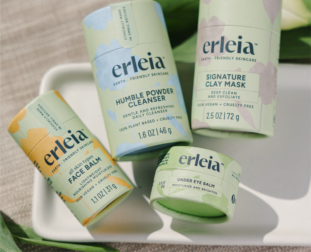 Erleia skincare products, including face balm, humble powder cleanser, signature clay mask, and under-eye balm, are displayed together in eco-friendly packaging, emphasizing their plant-based and cruelty-free formulations. They are sitting on a white tray with beige fabric and green leaves in the background.