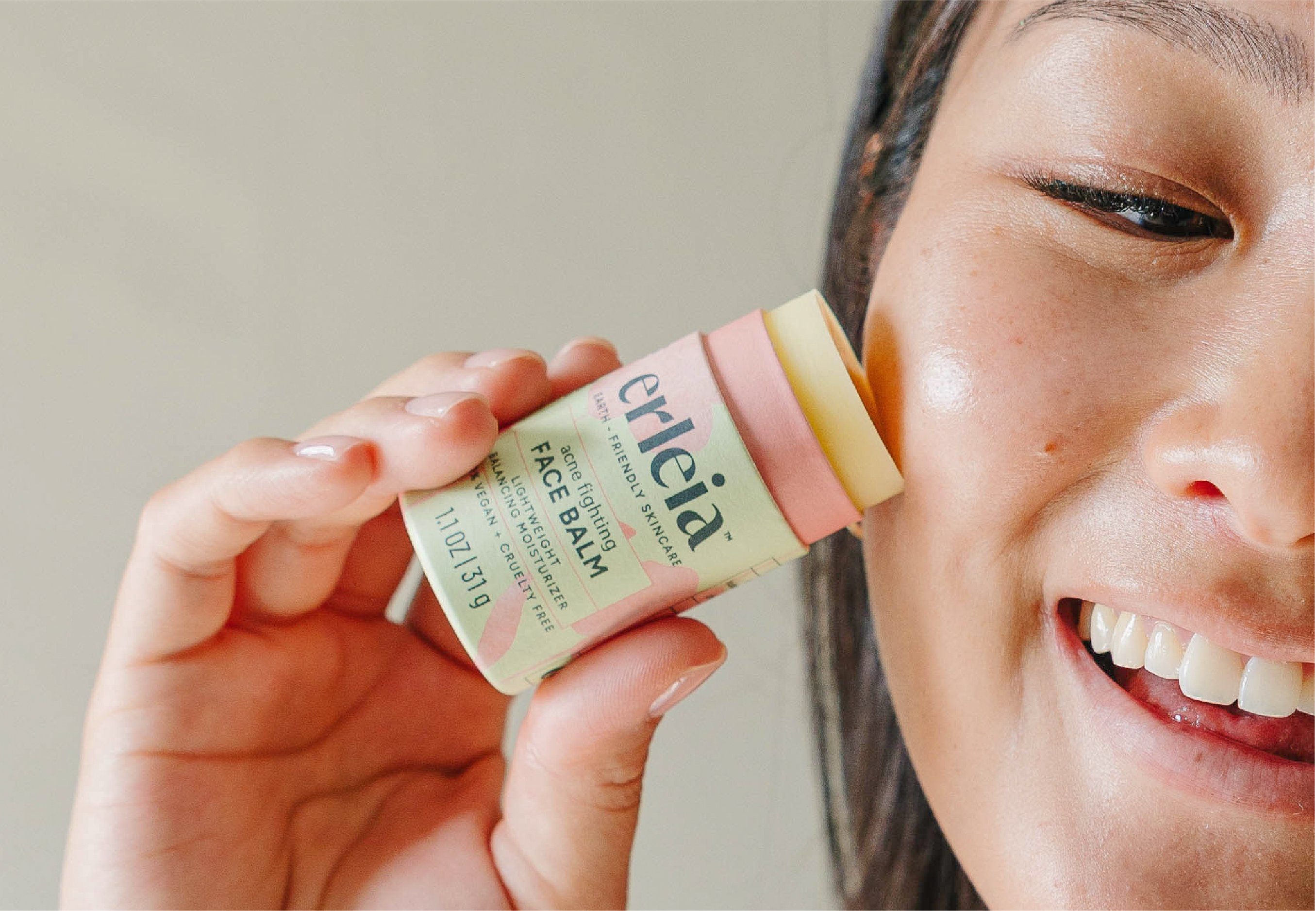 A smiling East Asian woman applies Erleia’s acne-fighting face balm to her cheek, showing the product’s eco-friendly packaging and highlighting its lightweight moisturizing benefits.