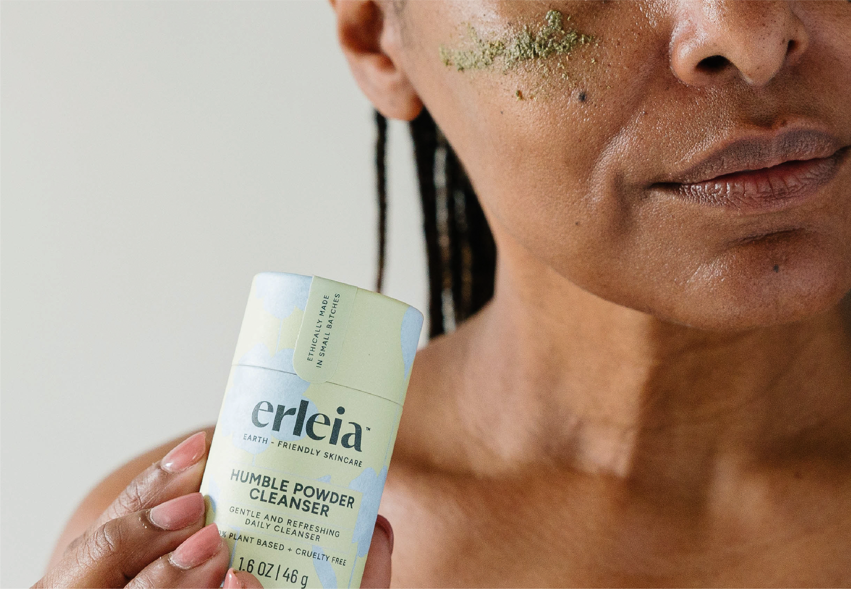 A Black woman with a green skincare product applied to their cheek holds Erleia’s Humble Powder Cleanser, highlighting its plant-based, cruelty-free formulation and eco-friendly packaging.