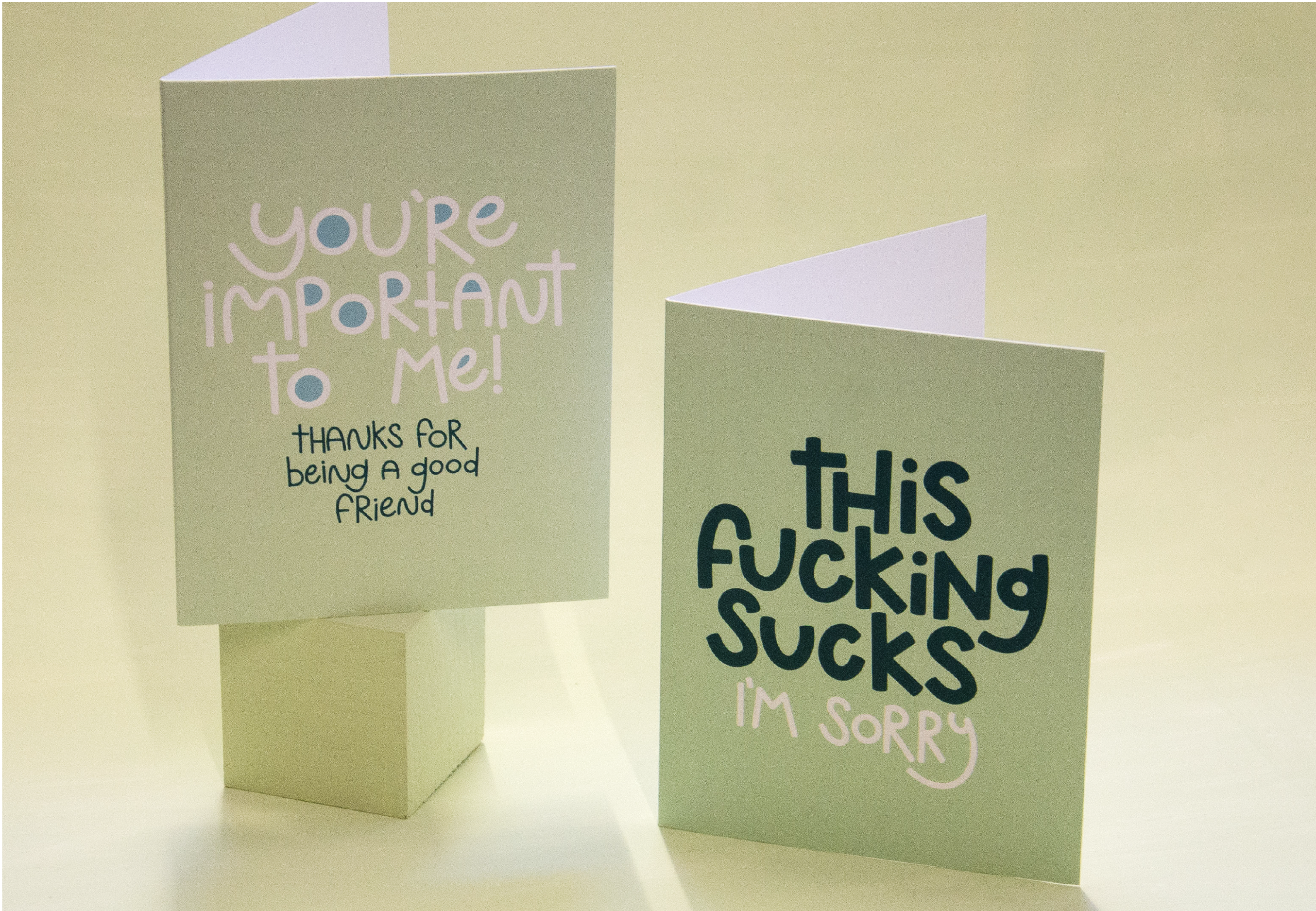 Two light green greeting cards are displayed on a light green backdrop, one reading "You're important to me! Thanks for being a good friend," and the other stating "This fucking sucks. I'm sorry."