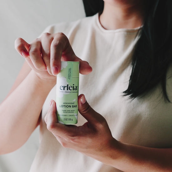 An East Asian woman wearing a beige top opens an Erleia unscented lotion bar, applies it to her arm and rubs it in with her hand.